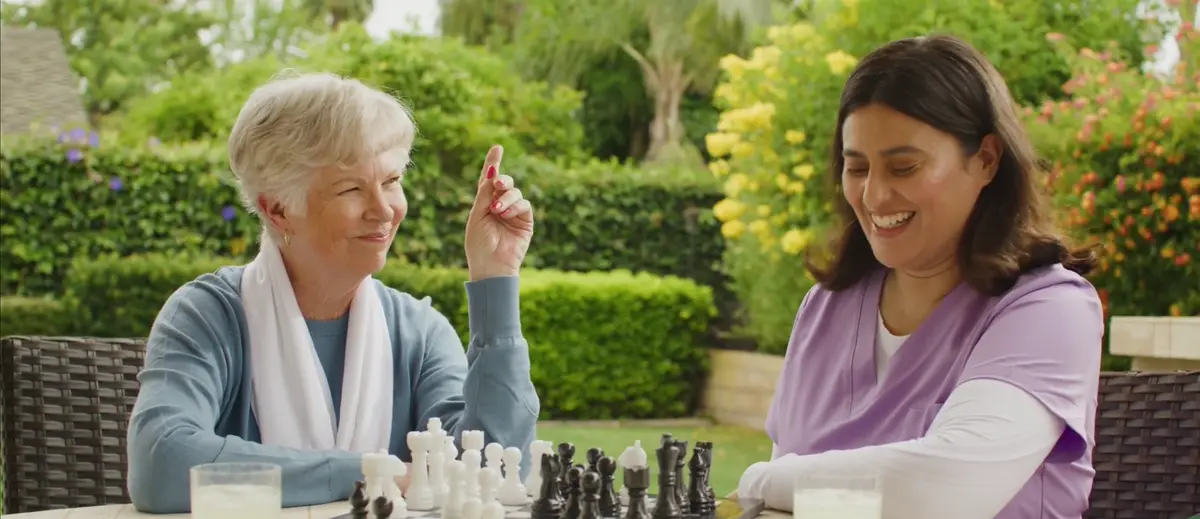 A caregiver and an older woman smiling over a game of chess in the garden
